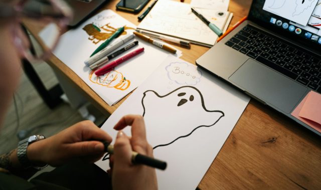 Pair of out-of-focus hands holding a felt tip pen over drawing of a white ghost in black felt tip outline with a speech bubble saying 'Bo-o-o' on white paper on a wooden desk next to a black laptop, more white paper with sketches and more pens.